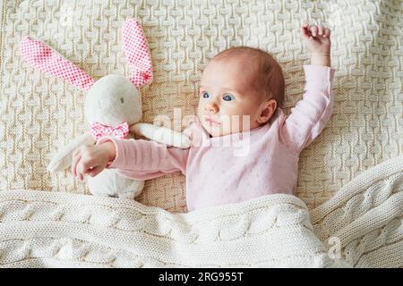 Une petite fille d'un mois couchée dans le lit sous la couverture tricotée avec son jouet, lapin en peluche rose Banque D'Images