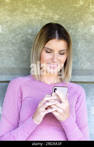 Une image de pur contentement - une femme blonde millénaire, vêtue d'une tenue de sport rose, s'appuie contre un mur de briques après une séance d'entraînement satisfaisante. Un sourire chaleureux orne son visage alors qu'elle se laisse tenter par des défilés et des messages bien mérités sur son smartphone, se prélassant dans l'arrière-lueur remplie d'endorphines de sa séance d'exercice. Post-Workout Bliss : Blonde Millennial Woman in Pink Sports Outfit appréciant son smartphone. Photo de haute qualité Banque D'Images