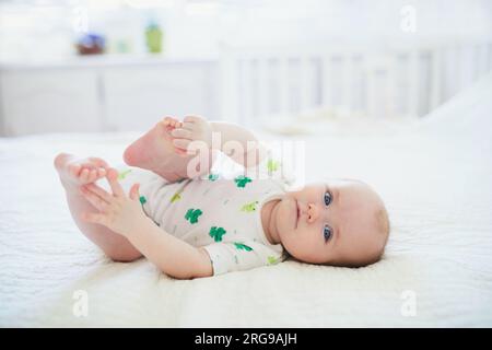 Mignonne petite fille allongée sur son dos et touchant ses pieds. Enfant heureux en bonne santé à la maison dans la pépinière Banque D'Images