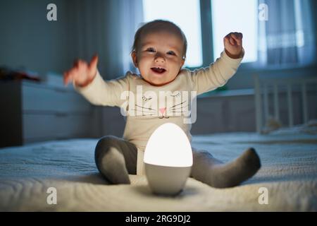 Adorable petite fille jouant avec la lampe de chevet dans la pépinière. Enfant heureux assis sur le lit avec veilleuse. Petit enfant à la maison le soir avant de dormir Banque D'Images