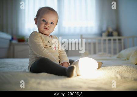 Adorable petite fille jouant avec la lampe de chevet dans la pépinière. Enfant heureux assis sur le lit avec veilleuse. Petit enfant à la maison le soir avant de dormir Banque D'Images