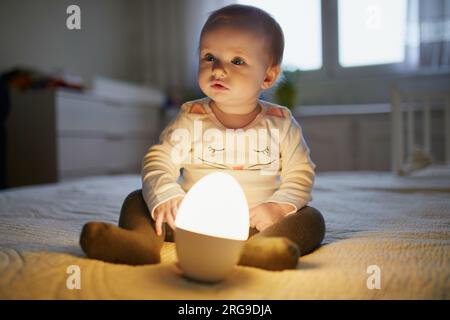 Adorable petite fille jouant avec la lampe de chevet dans la pépinière. Enfant heureux assis sur le lit avec veilleuse. Petit enfant à la maison le soir avant de dormir Banque D'Images