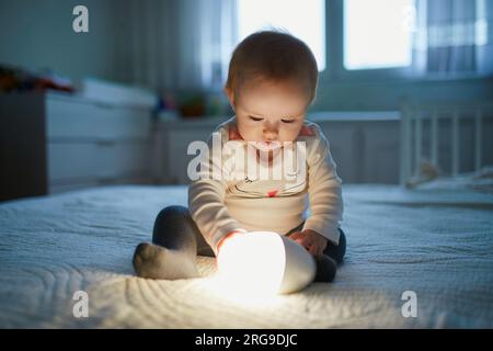 Adorable petite fille jouant avec la lampe de chevet dans la pépinière. Enfant heureux assis sur le lit avec veilleuse. Petit enfant à la maison le soir avant de dormir Banque D'Images