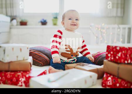 Heureuse petite fille de bébé portant un pull chaud de vacances ouvrant des cadeaux de Noël sur son premier Noël. Fêter Noël avec les enfants à la maison Banque D'Images