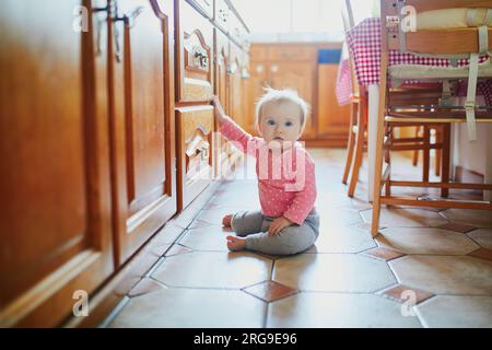 Petite fille assise sur le sol dans la cuisine. Petit enfant rampant à la maison et explorant les choses autour d'elle Banque D'Images