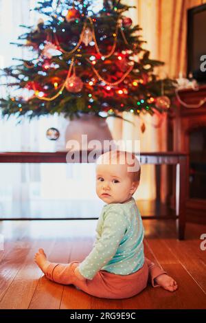 Petite fille assise sur le sol sous l'arbre de Noël. Petit enfant rampant à la maison et explorant les choses autour d'elle. Vacances avec petits enfants Banque D'Images