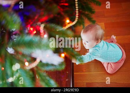 Petite fille assise sur le sol sous l'arbre de Noël. Petit enfant rampant à la maison et explorant les choses autour d'elle. Vacances avec petits enfants Banque D'Images