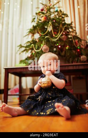 Bébé fille assise dans une belle robe sur le sol sous l'arbre de Noël. Petit enfant rampant à la maison et explorant les choses autour d'elle. Vacances avec l Banque D'Images