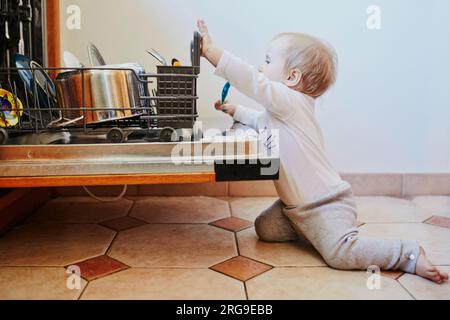 Petit enfant aidant à décharger le lave-vaisselle. Petite fille assise sur le sol dans la cuisine. Petit enfant à la maison Banque D'Images