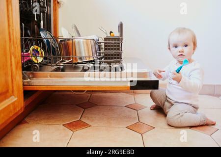 Petit enfant aidant à décharger le lave-vaisselle. Petite fille assise sur le sol dans la cuisine. Petit enfant à la maison Banque D'Images