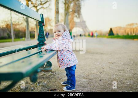 Une fille d'un an debout à côté du banc près de la tour Eiffel. Tout-petit apprenant à marcher. Adorable enfant marchant à Paris, France. Voyager avec Banque D'Images