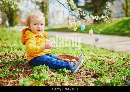 Mignonne petite fille d'un an jouant à la chasse aux œufs à Pâques. Tout-petit assis sur l'herbe sous le pommier en pleine floraison décoré avec des œufs colorés. Li Banque D'Images