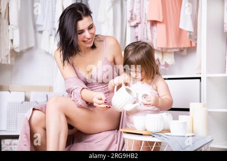 Jeune belle brune en body rose joue avec sa petite fille dans un dressing lumineux. Banque D'Images