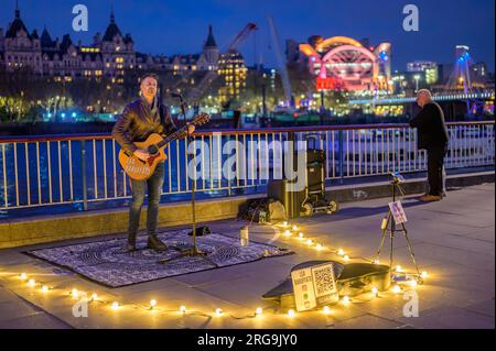 LONDRES - 22 avril 2023 : nuit enchanteresse : Busker divertit les touristes sur Southbank, des lumières chaudes contre un ciel bleu profond, la station Charing Cross brille Ora Banque D'Images