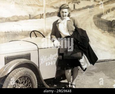 Une jeune femme sur le front de mer de Blackpool pose pour une photo « Sunny Snaps » aux côtés d'une petite voiture de sport « prop » devant un décor peint représentant une scène de campagne. Banque D'Images