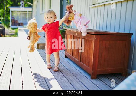 Adorable petite fille en robe rouge jouant avec poupée et ours en peluche à l'extérieur sur une journée d'été ensoleillée. Petit enfant s'amusant. Gamin jouant aux jouets en peluche Banque D'Images