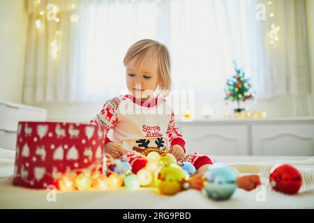 Heureuse petite fille en bas âge portant un pyjama et jouant avec des décorations de Noël sur le lit. Célébrer les vacances saisonnières avec les enfants à la maison Banque D'Images