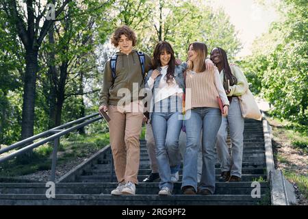 Un groupe d'adolescents divers avec des sacs à dos et des cahiers conversent alors qu'ils descendent les escaliers du parc, capturés de la taille vers le haut. S'engager dans un con animé Banque D'Images