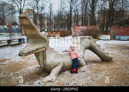 Adorable petite fille en bas âge dans le parc sur un jour de printemps ou d'hiver. Enfant sur l'aire de jeux s'amusant avec statue de crocodile en pierre. Activités de plein air pour les enfants Banque D'Images