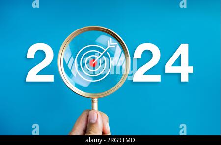 Bonne année 2024 avec bannière de concept d'entreprise. Le grand numéro blanc de 2024 ans avec l'icône de fléchette cible à l'intérieur de la loupe dorée en main sur b Banque D'Images