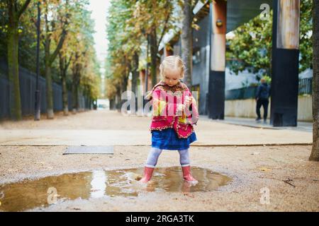 Enfant portant des bottes de pluie rouges et sautant dans la flaque d'eau un jour d'automne. Adorable petite fille s'amusant avec de l'eau et de la boue dans le parc un jour de pluie. Extérieur Banque D'Images