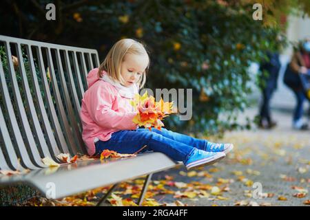 Adorable petite fille assise sur le banc et rassemblant les feuilles tombées dans le parc d'automne. Enfant heureux profitant de la journée d'automne à Paris, France. Activités de plein air Banque D'Images