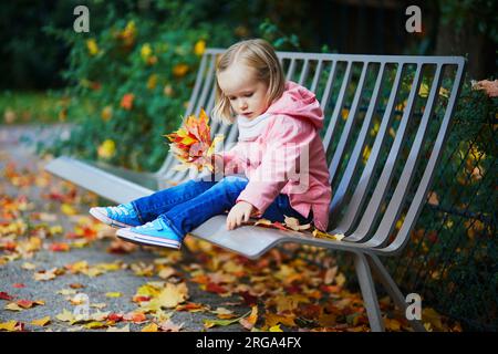 Adorable petite fille assise sur le banc et rassemblant les feuilles tombées dans le parc d'automne. Enfant heureux profitant de la journée d'automne à Paris, France. Activités de plein air Banque D'Images