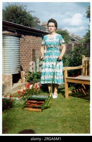 Femme d'âge moyen élégamment habillée debout avec fierté (si légèrement envolez-vous) dans un jardin de banlieue avec sa tondeuse. Banque D'Images