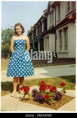 Une jolie jeune femme dans une robe bleue et à pois dans un jardin de banlieue britannique impeccable - à côté d'un petit lit en forme de losange fleuri contenant des œillets rouges - septembre 1959 Banque D'Images