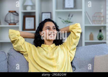 Jeune belle femme hispanique se détendant à la maison de près, femme avec les mains derrière la tête souriant et regardant par la fenêtre, rêvant assis sur le canapé dans le salon à la maison. Banque D'Images