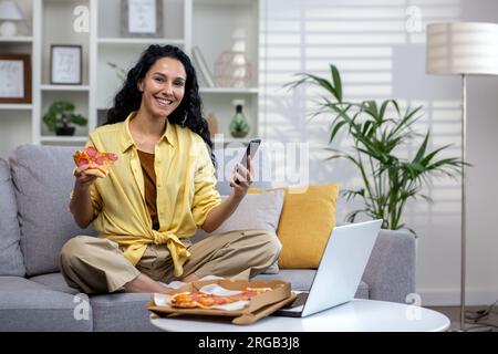 Portrait de la femme cliente heureuse de livraison de nourriture en ligne, femme hispanique souriante et regardant la caméra, en utilisant l'application sur le téléphone pour commander la livraison de pizza. Banque D'Images