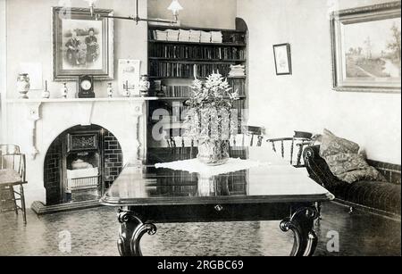 Élégante salle de réception édouardienne avec table en bois poli, chaise longue, grand portrait photographique de famille au-dessus de la cheminée et étagères soignées aux volumes en cuir. Banque D'Images