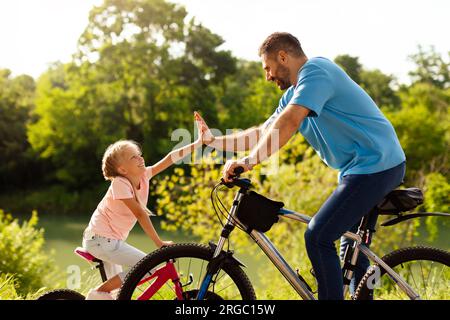 Père et sa fille donnant haut cinq tout en faisant du vélo dans le parc ensoleillé, profiter de passer du temps libre ensemble, vue de côté Banque D'Images