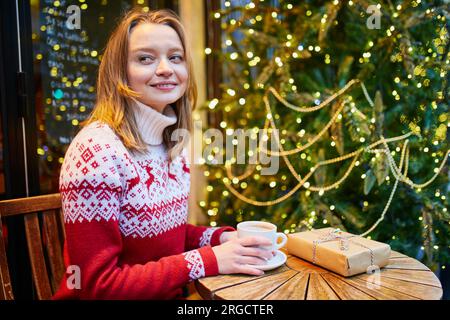 Jeune fille joyeuse dans le pull de vacances boire du café ou du chocolat chaud dans le café décoré pour Noël et déballer le cadeau de Noël Banque D'Images