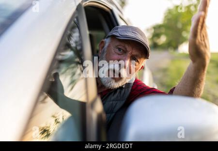 Homme senior conduisant une voiture, regardant par la fenêtre latérale et se disputant avec quelqu'un Banque D'Images