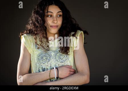 Jolie jeune fille confuse debout avec les bras croisés dans le studio avec fond sombre Banque D'Images