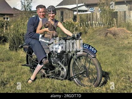 Un couple assis sur une moto dans un jardin. La femme tient un spaniel sur ses genoux. Banque D'Images