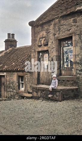 Scène de rue dans les Highlands écossais, avec une petite fille assise sur un pas de pierre. Une sculpture en pierre d'une balance porte l'inscription: Dieu bénisse le juste. Banque D'Images