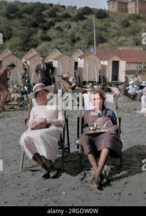 Deux jeunes femmes assises sur une plage dans des transats, avec des cabines de bain en arrière-plan. Banque D'Images