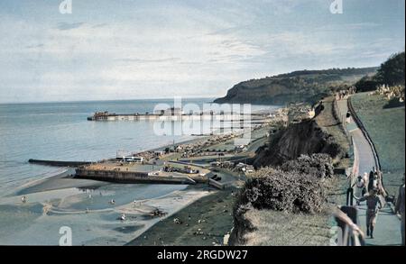 Une station balnéaire non identifiée avec un quai et une jetée, et des personnes marchant le long d'un chemin de falaise sur la droite. Banque D'Images