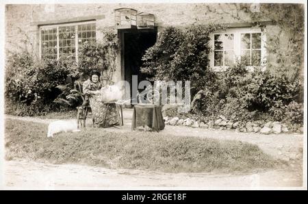 Une dame âgée (entourée de ses nombreux animaux) dentelle faisant à l'extérieur de son charmant chalet rural. Une petite roue qui tourne se tient à côté. Banque D'Images
