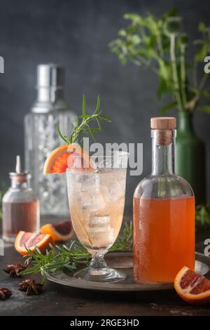 Cocktail clair sur glace avec sirop d'orange sanguine et club soda sur glace en verre vintage avec bouteille de sirop, garniture d'agrumes et de romarin Banque D'Images