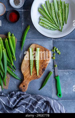 Tranches d'okra en forme de frites dans un bol blanc et une gousse d'okra coupée en deux dans le sens de la longueur sur une planche à découper, un couteau sur le côté. Banque D'Images