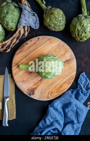 Un artichaut sur une planche à découper ronde en bois, un couteau et plus d'artichauts accompagnent. Banque D'Images