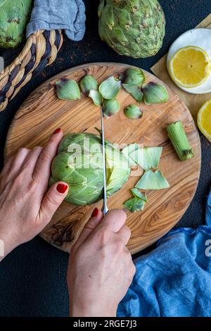 Mains de femme coupant le dessus d'un artichaut sur une planche de bois. Banque D'Images