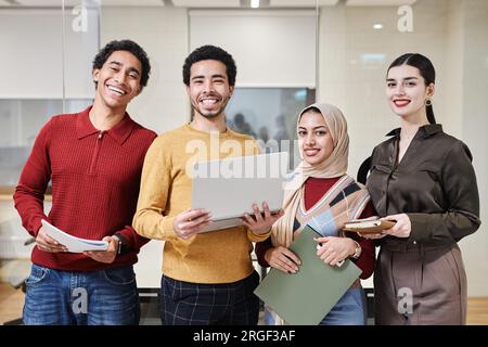 Portrait à la taille de l'équipe d'affaires du Moyen-Orient debout dans un bureau moderne et souriant à la caméra tenant joyeusement les ordinateurs et les documents Banque D'Images