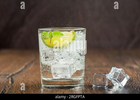 Eau minérale rafraîchissante dans un verre avec glaçons, feuilles de menthe et tranche de citron vert. Vaporiser sur un verre d'eau. Fond en bois. Banque D'Images