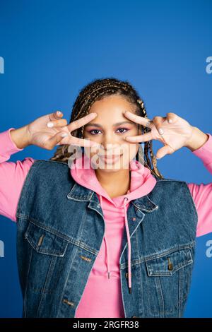 femme afro-américaine attrayante et heureuse sur fond bleu, gilet en denim, montrant le signe v. Banque D'Images