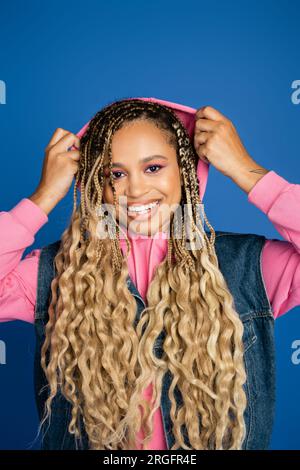 femme à peau foncée positive portant une capuche rose sur la tête, regardant la caméra sur fond bleu Banque D'Images