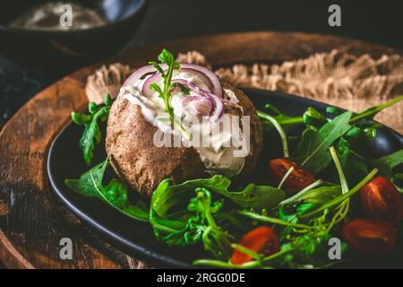 Pommes de terre cuites avec de la crème sure, des oignons et des feuilles de roquette avec des tomatos sur une assiette noire sur une table en bois rustique Banque D'Images